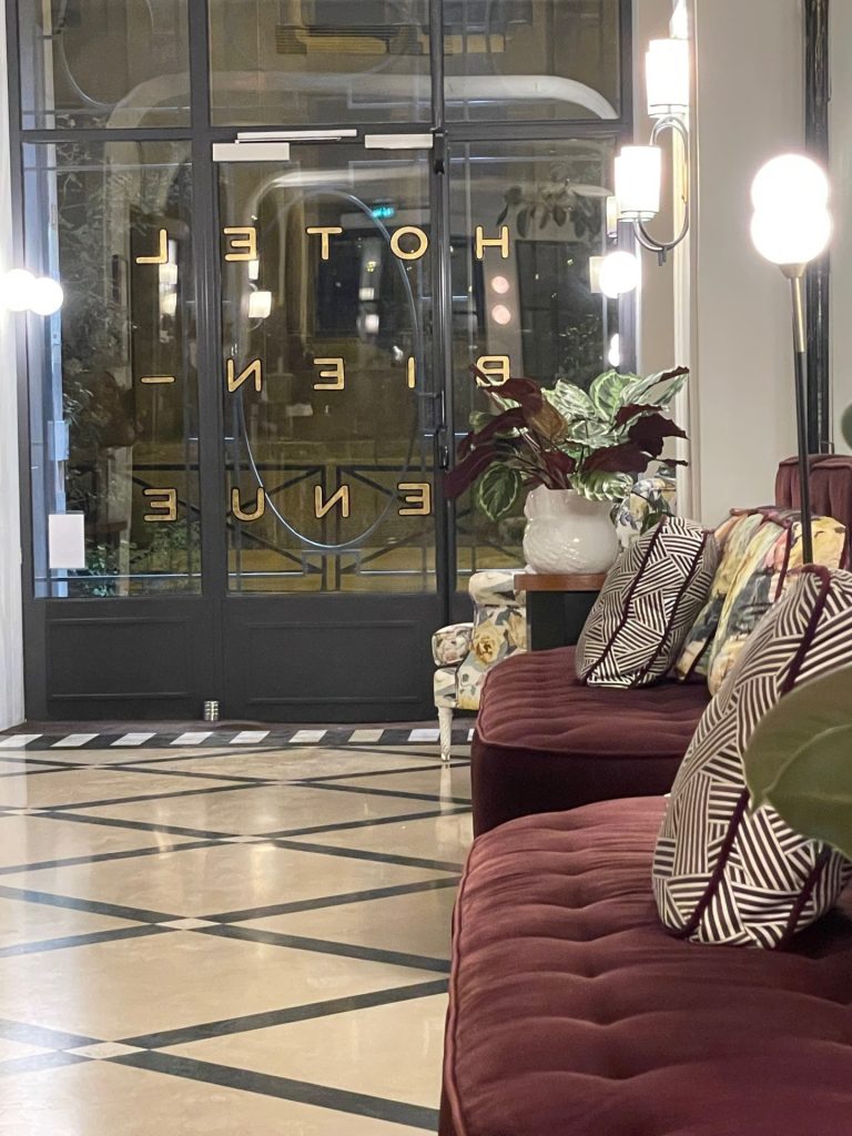 View of lobby hall with velvet couches leading to the glass entry door with the name of the hotel in gold letters.